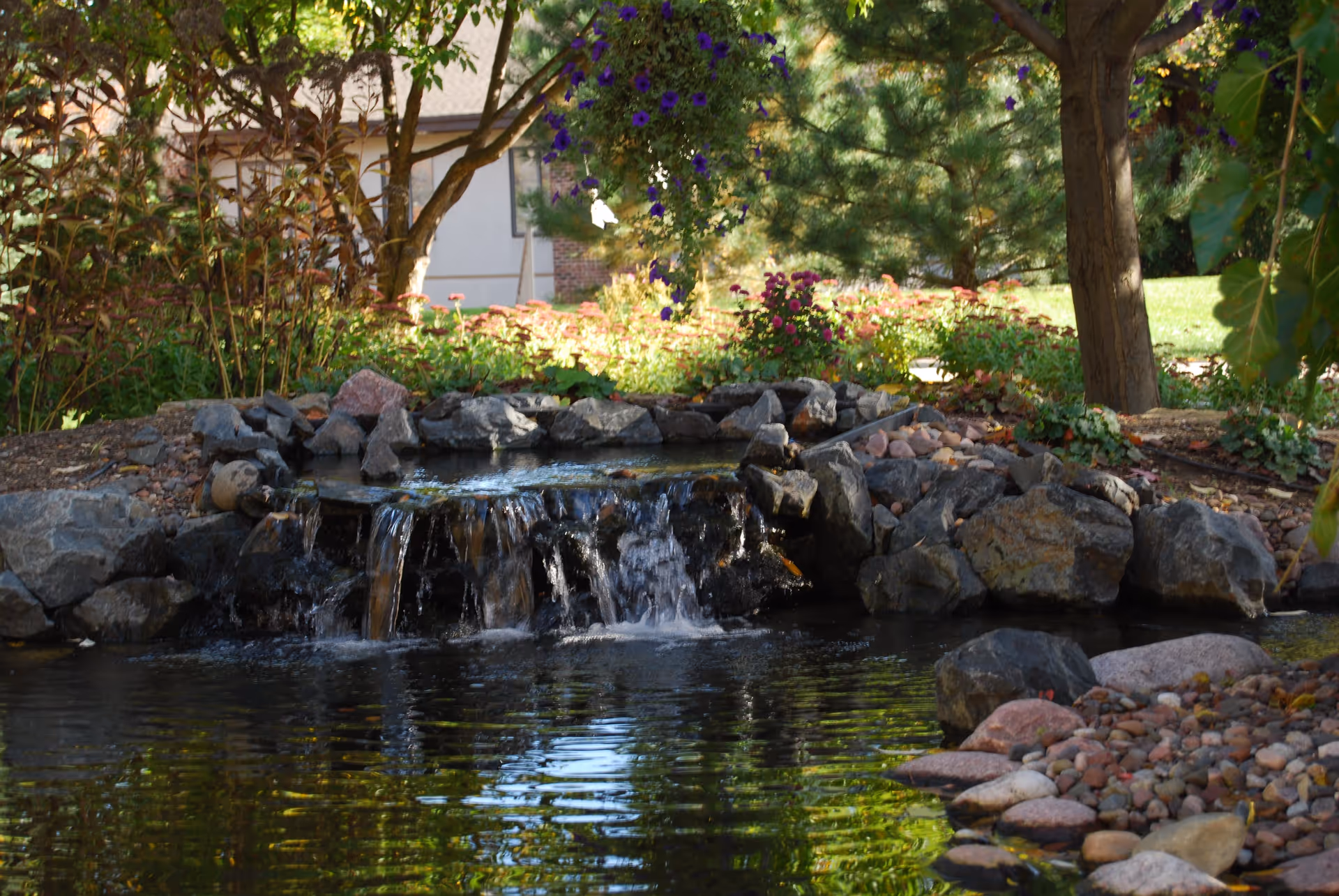 A small landscaped garden pond with a waterfall flowing over rocks surrounded by trees, flowers, and greenery with a building partially visible in the background.