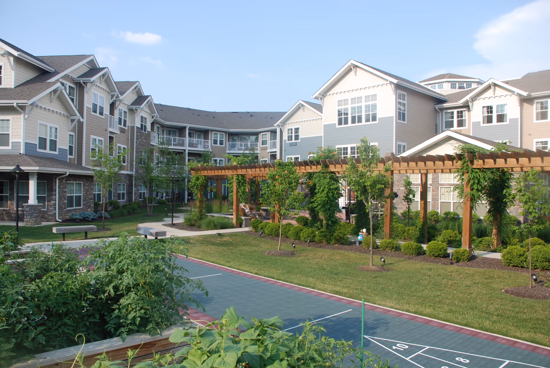 Outdoor view of Northridge Gracious Retirement Living showing a landscaped garden area with a wooden pergola, green shrubs, small trees, and a shuffleboard court in the foreground. The multi-story residential building with balconies and large windows surrounds the garden area under a clear blue sky.