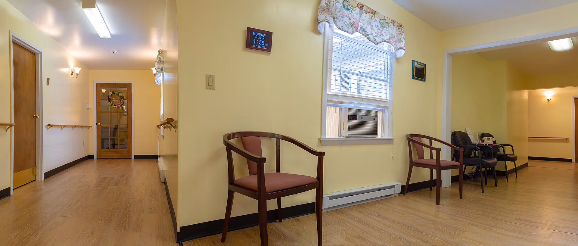 Interior hallway of Plymouth Manor Personal Care Center with light yellow walls, wooden flooring, several chairs along the walls, a window with floral valance and an air conditioning unit, and a door with a wreath at the end of the hallway.
