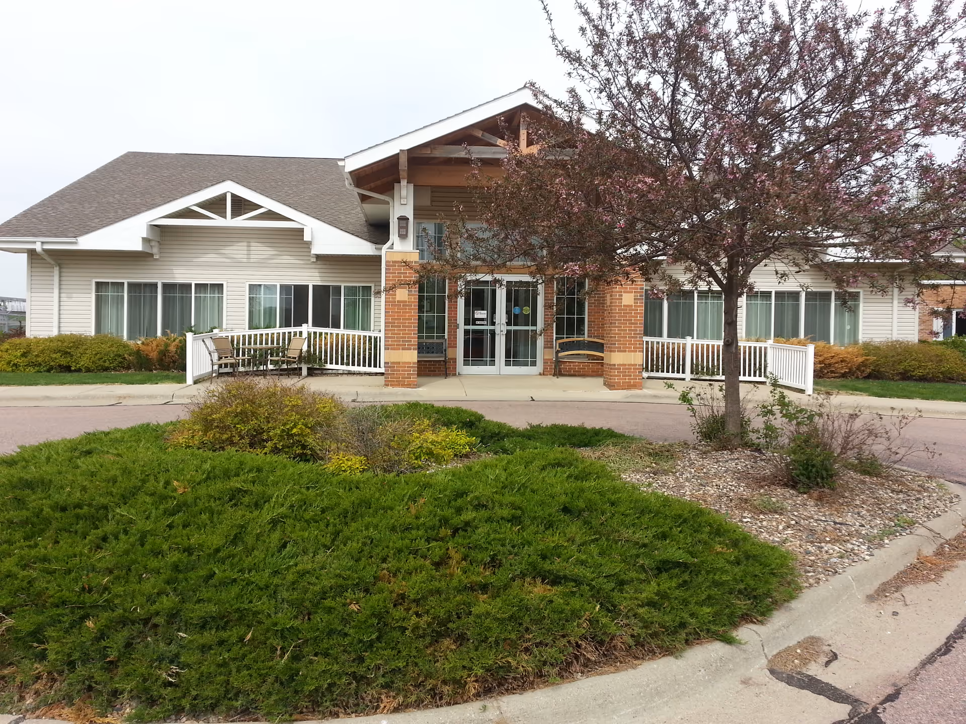 Exterior front entrance of Countryside Health Care Center with a covered porch supported by brick columns, a glass double door entrance, a tree with pink blossoms, green shrubs, and a paved driveway.