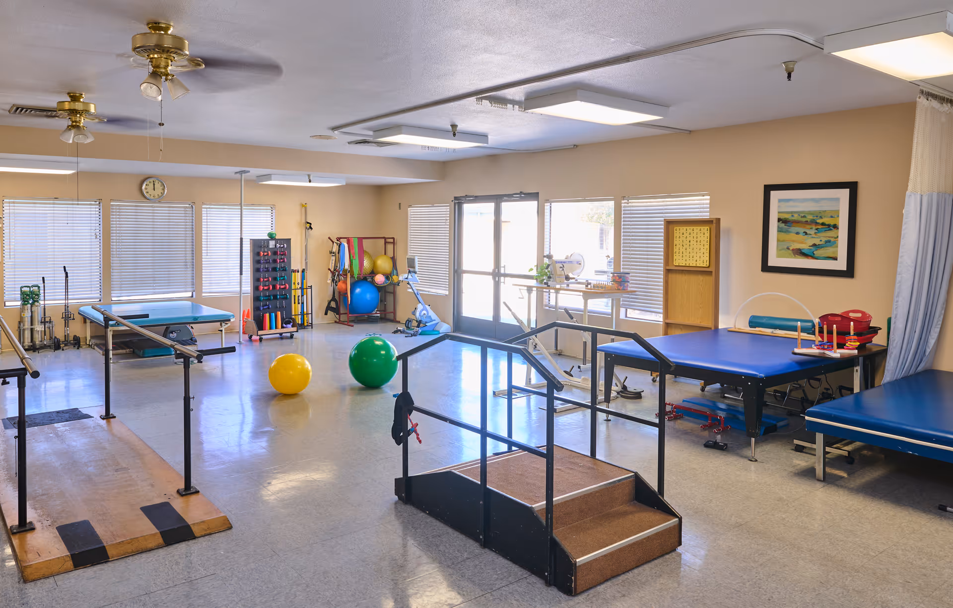 Rehabilitation therapy room with parallel bars, exercise balls, treatment tables, and a small practice staircase.