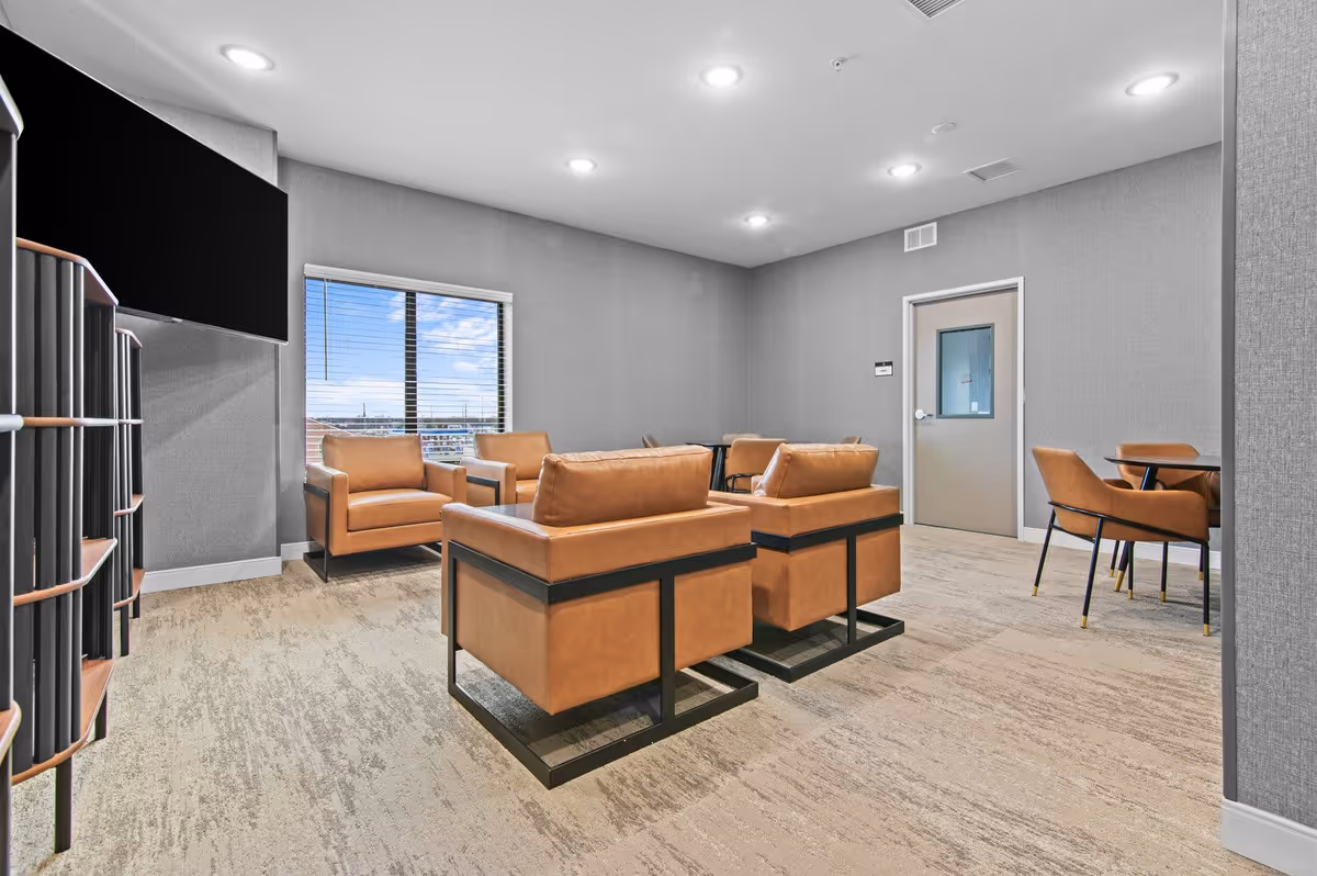A modern lounge area with four tan leather armchairs arranged around a small table. There is a large flat-screen TV mounted on the wall to the left and a window with blinds showing a view of the outside. To the right, there is a small table with two matching tan chairs. The room has gray walls, recessed ceiling lights, and beige carpet flooring.