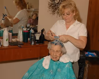 An elderly woman sitting in a salon chair with a green cape around her shoulders while a hairstylist applies hair rollers to her hair. Various hair care products and tools are visible on the counter behind them, along with a mirror reflecting the scene.