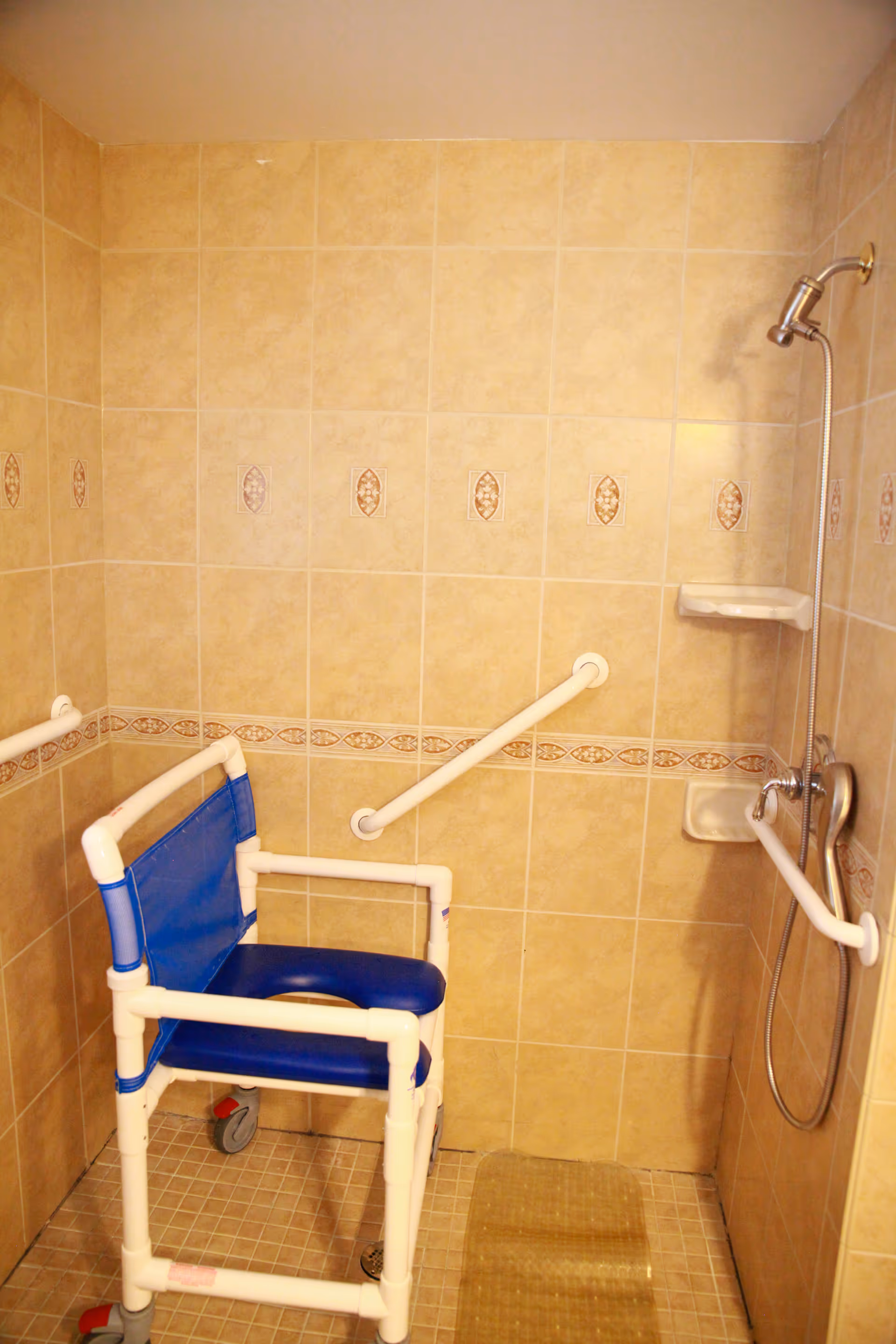 Tiled walk-in shower with beige tiles, multiple grab bars, a handheld showerhead, and a blue wheeled shower chair.