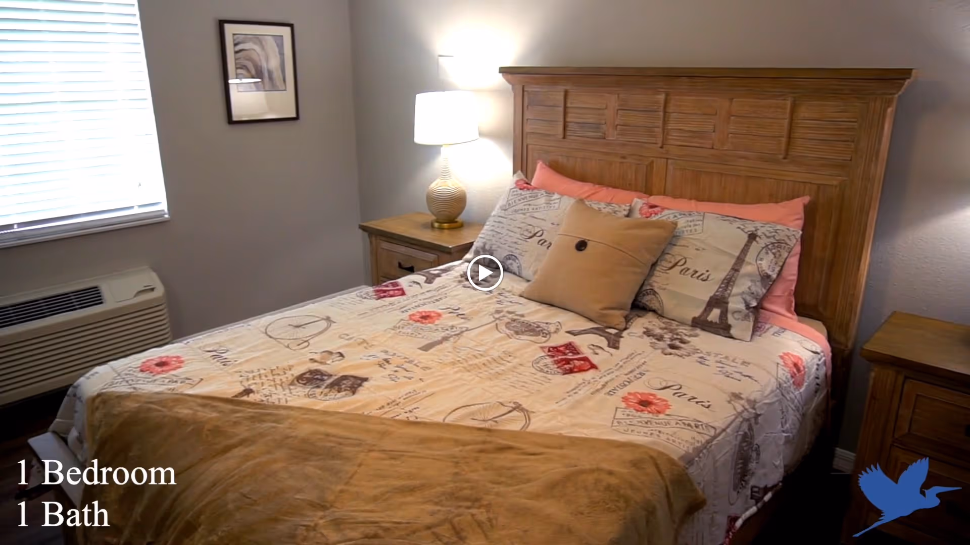 A cozy bedroom with a wooden headboard bed dressed in Paris-themed bedding, including pillows and a beige throw blanket. There is a wooden nightstand with a lamp on the right side of the bed and another nightstand partially visible on the left. A window with blinds and an air conditioning unit is on the left wall, and a framed picture hangs above the nightstand.