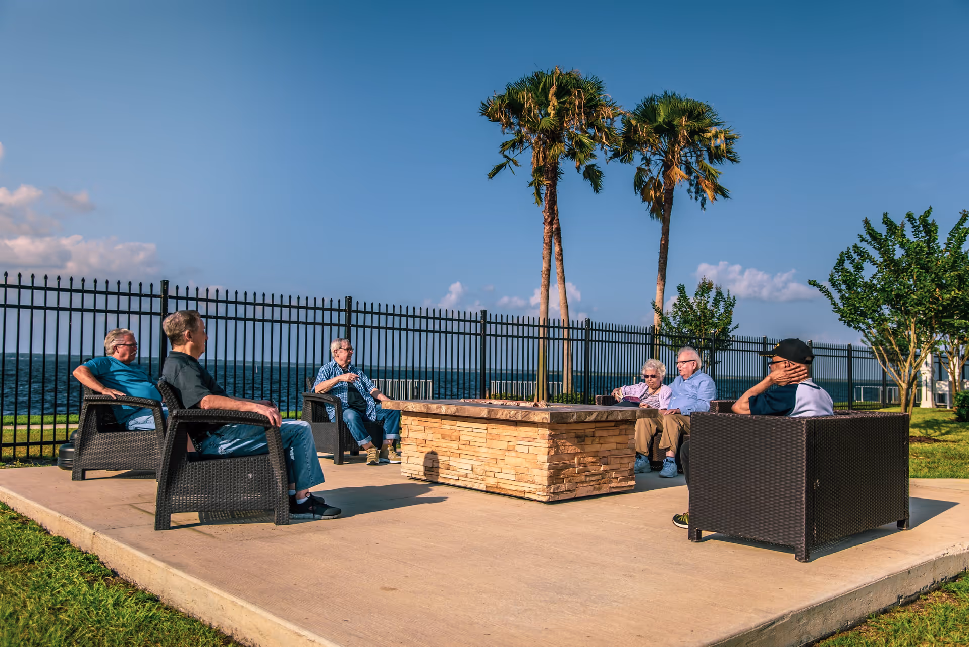 A group of six elderly people sitting outdoors in a semi-circle around a stone fire pit on a concrete patio. They are seated on dark wicker chairs with a black metal fence, palm trees, green grass, and a body of water visible in the background under a clear blue sky.