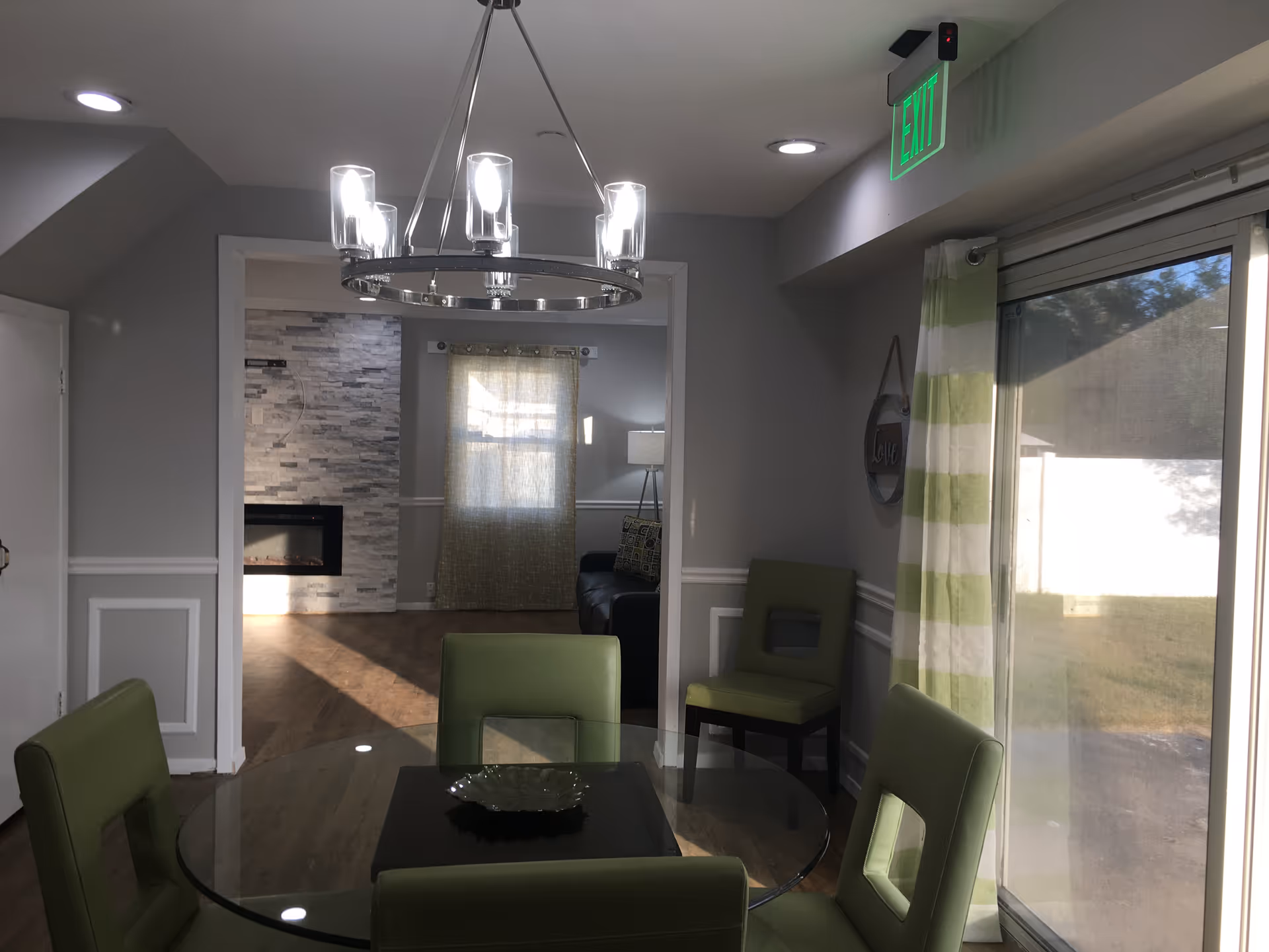 Sunlit dining area with a glass table and green chairs, a modern chandelier, sliding glass door, and a view into a living room with a fireplace.