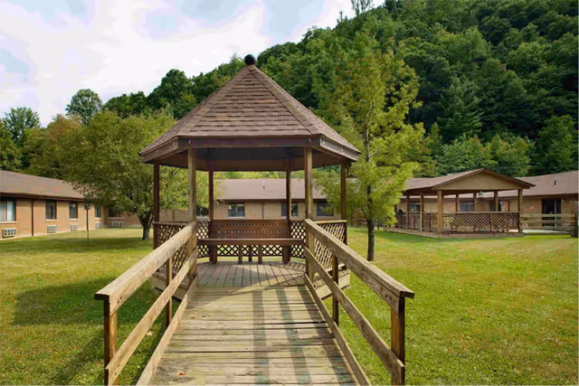 A wooden gazebo with a shingled roof at the end of a wooden walkway, surrounded by green grass and trees, with single-story brick buildings in the background and a forested hill behind them.