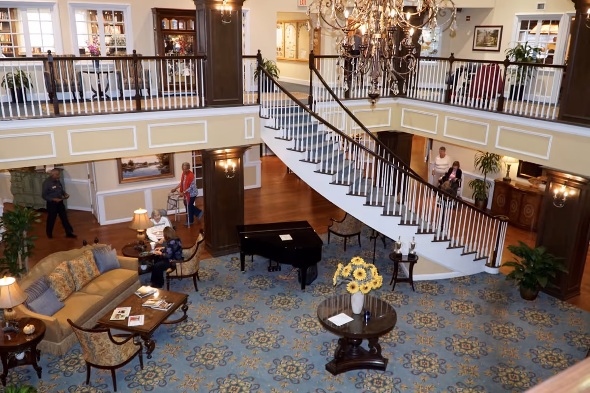 Spacious senior living facility common area with a grand staircase, chandelier, patterned carpet, and seating arrangements including a sofa and chairs. Several seniors are seen interacting and walking around the area. A black grand piano and a round table with a vase of yellow flowers are also visible.