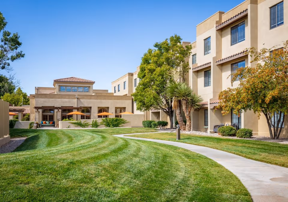 Outdoor view of a senior living facility with a well-maintained green lawn, a curved concrete walkway, trees, and a multi-story beige building with windows. There are orange umbrellas and outdoor seating visible near the building under a clear blue sky.