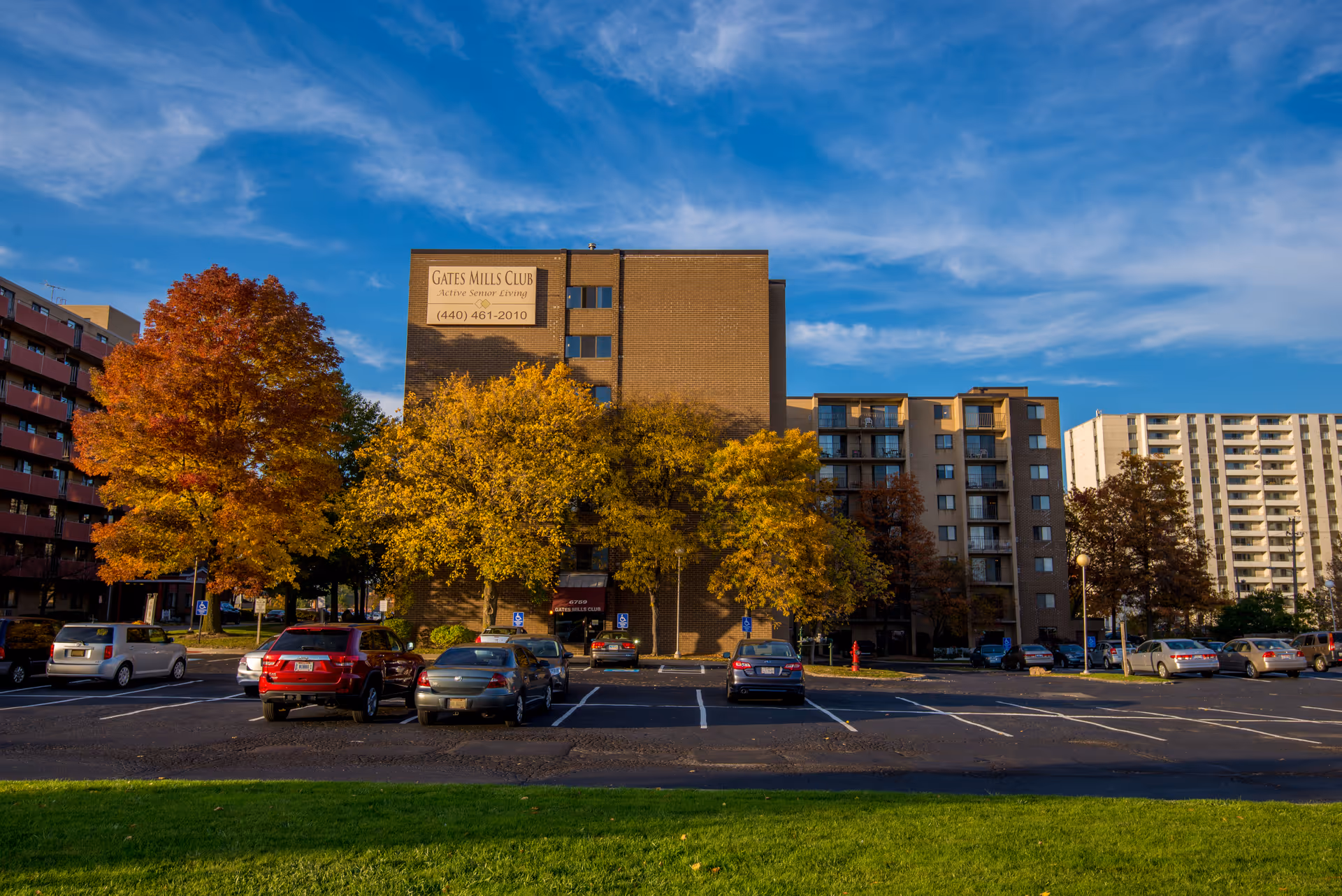 Front view of the Gates Mills Club senior living building with a parking lot and autumn trees under a blue sky.