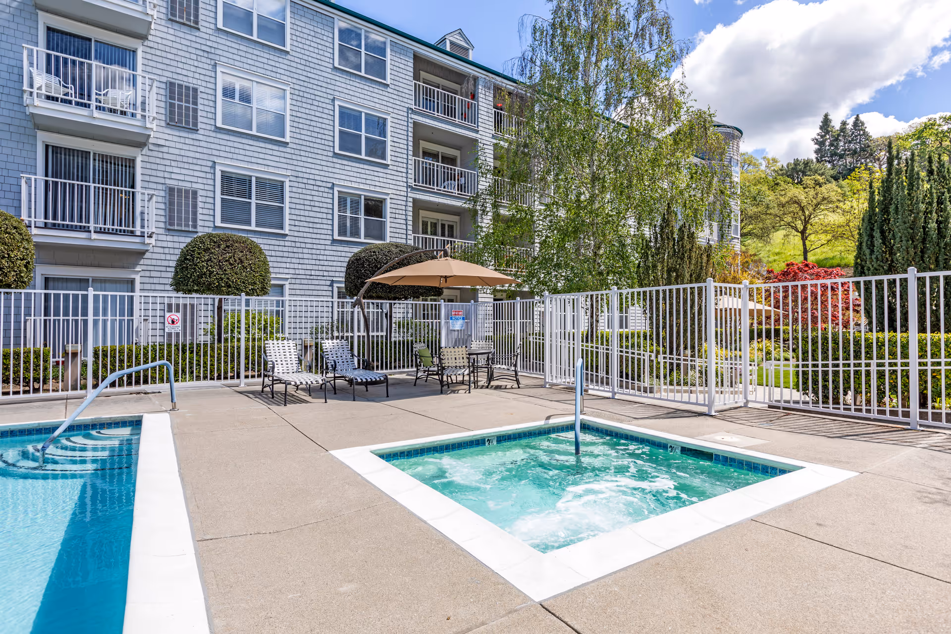 Outdoor pool and hot tub area at a senior living facility named Byron Park, with lounge chairs, a table with an umbrella, and a multi-story building with balconies in the background surrounded by trees and landscaping.