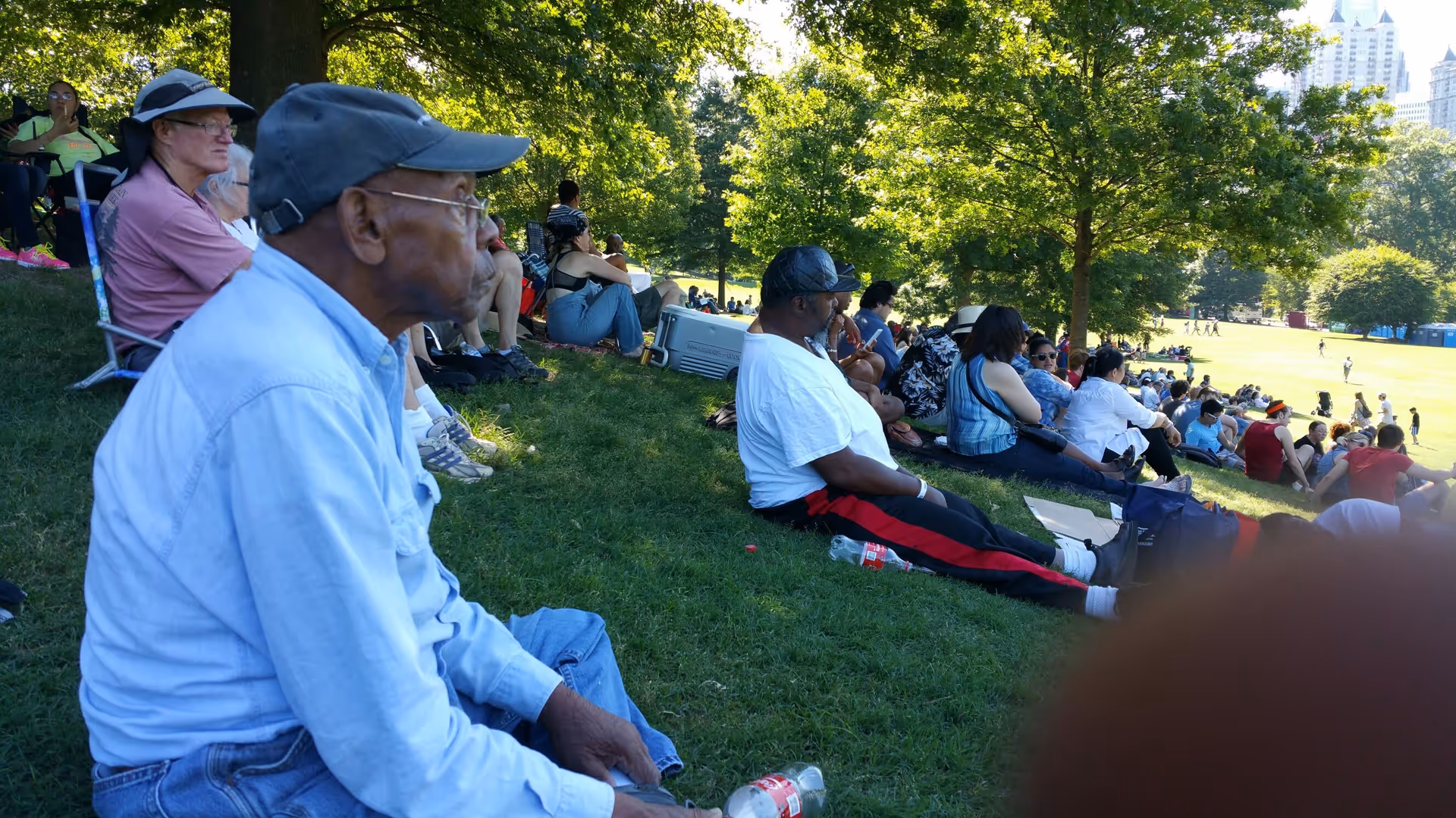 A group of people sitting on a grassy hill in a park, watching an event or activity taking place on a field. The scene is shaded by trees with sunlight filtering through, and some people are seated on portable chairs while others sit directly on the grass.