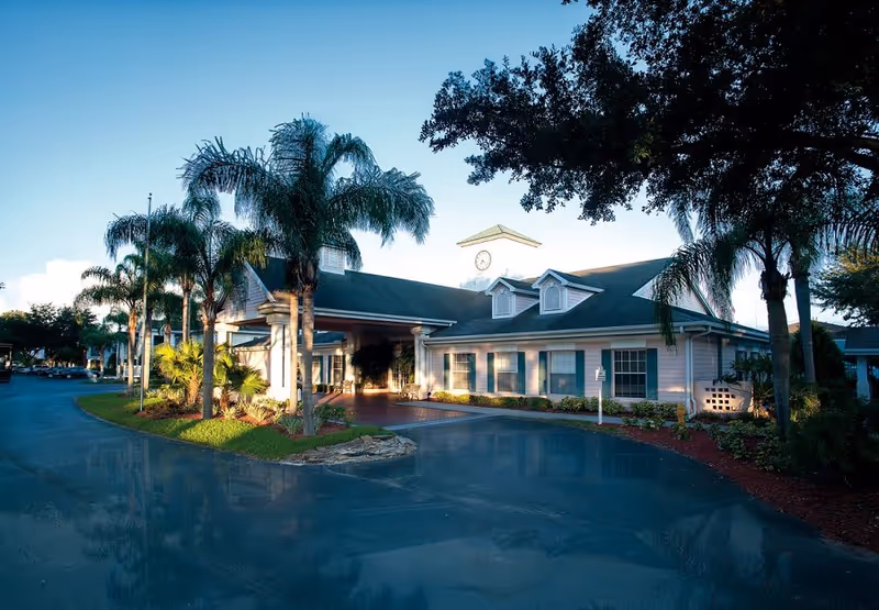 Exterior view of a senior living facility building with a green roof, white walls, and a clock tower. The building is surrounded by palm trees and landscaping, with a paved driveway in front.