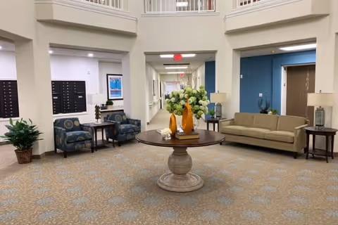 A spacious and well-lit common area in a retirement living facility featuring a round wooden table with decorative vases and flowers in the center. The room has patterned carpet flooring, a beige sofa, two patterned armchairs with a small table between them, potted plants, and a hallway leading to other rooms. The walls are painted in neutral tones with some blue accent walls, and there are ceiling lights providing illumination.