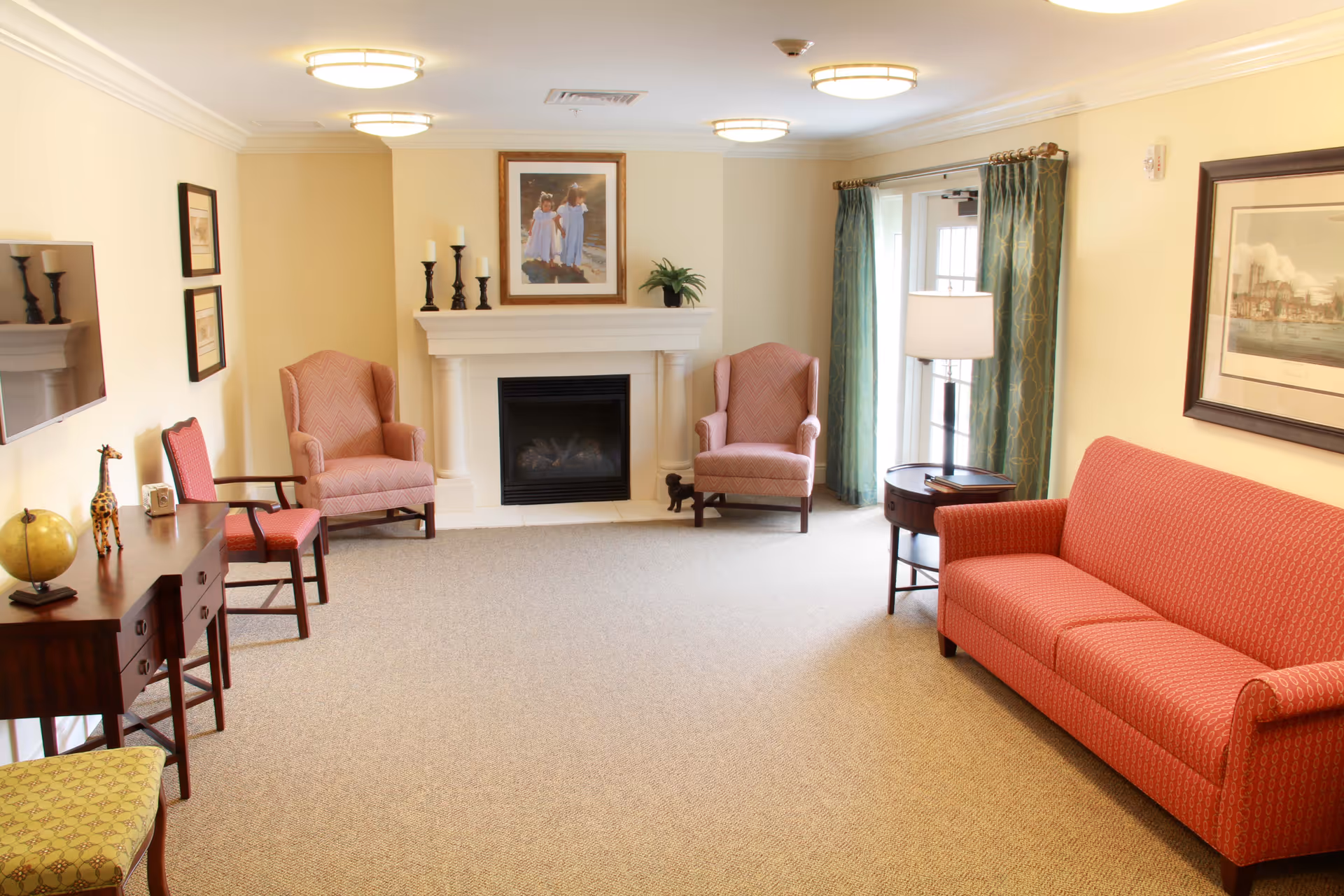 A cozy senior living room with a red patterned sofa, two matching armchairs, a wooden side table with a lamp, a fireplace with candles and a framed picture above it, a TV mounted on the wall, and large windows with green curtains allowing natural light to enter.