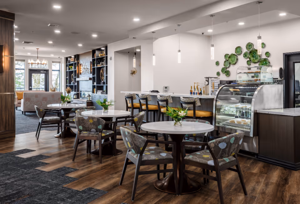 Interior view of a modern dining and lounge area in Boone Ridge Retirement Community featuring round tables with floral-patterned chairs, a counter with bar stools, a display case with desserts, decorative green wall art, and a cozy seating area with sofas near large windows.