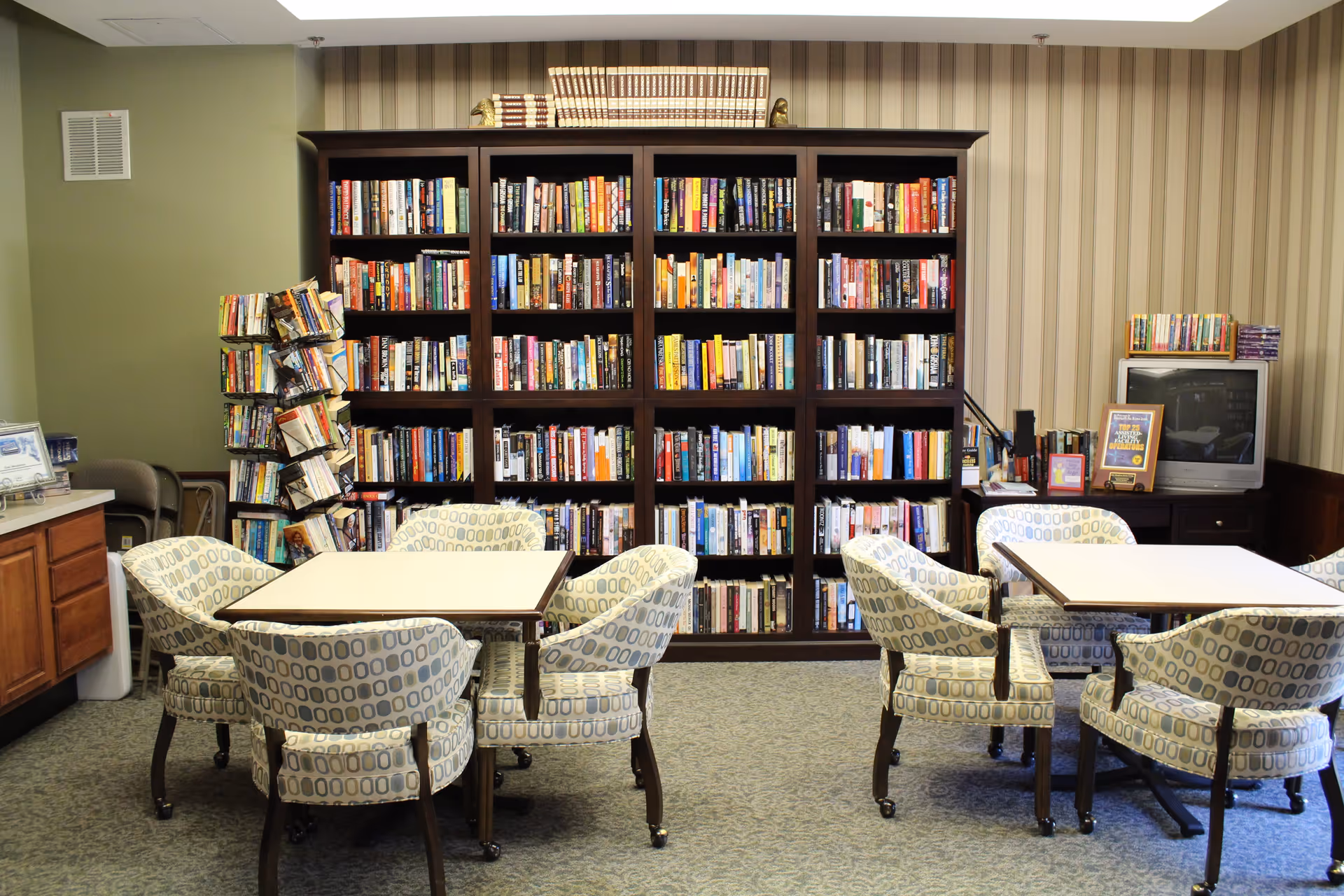 A cozy reading room in Oak Meadows Senior Living featuring two square tables each surrounded by four cushioned chairs with patterned upholstery. Behind the tables is a large dark wooden bookshelf filled with numerous books. To the left, there is a rotating book rack with more books. On the right side, a small TV sits on a cabinet with additional books and framed awards or certificates. The walls have a combination of green paint and striped wallpaper.