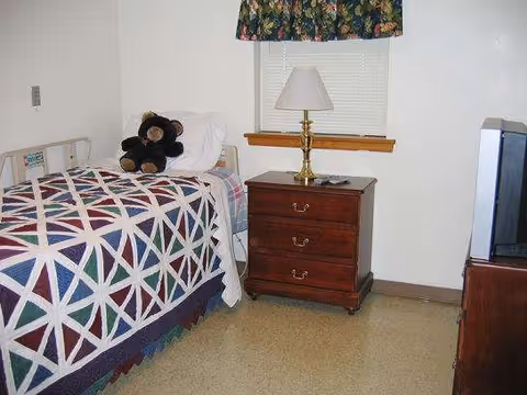 A simple bedroom with a quilted bed, nightstand, lamp, and a teddy bear.
