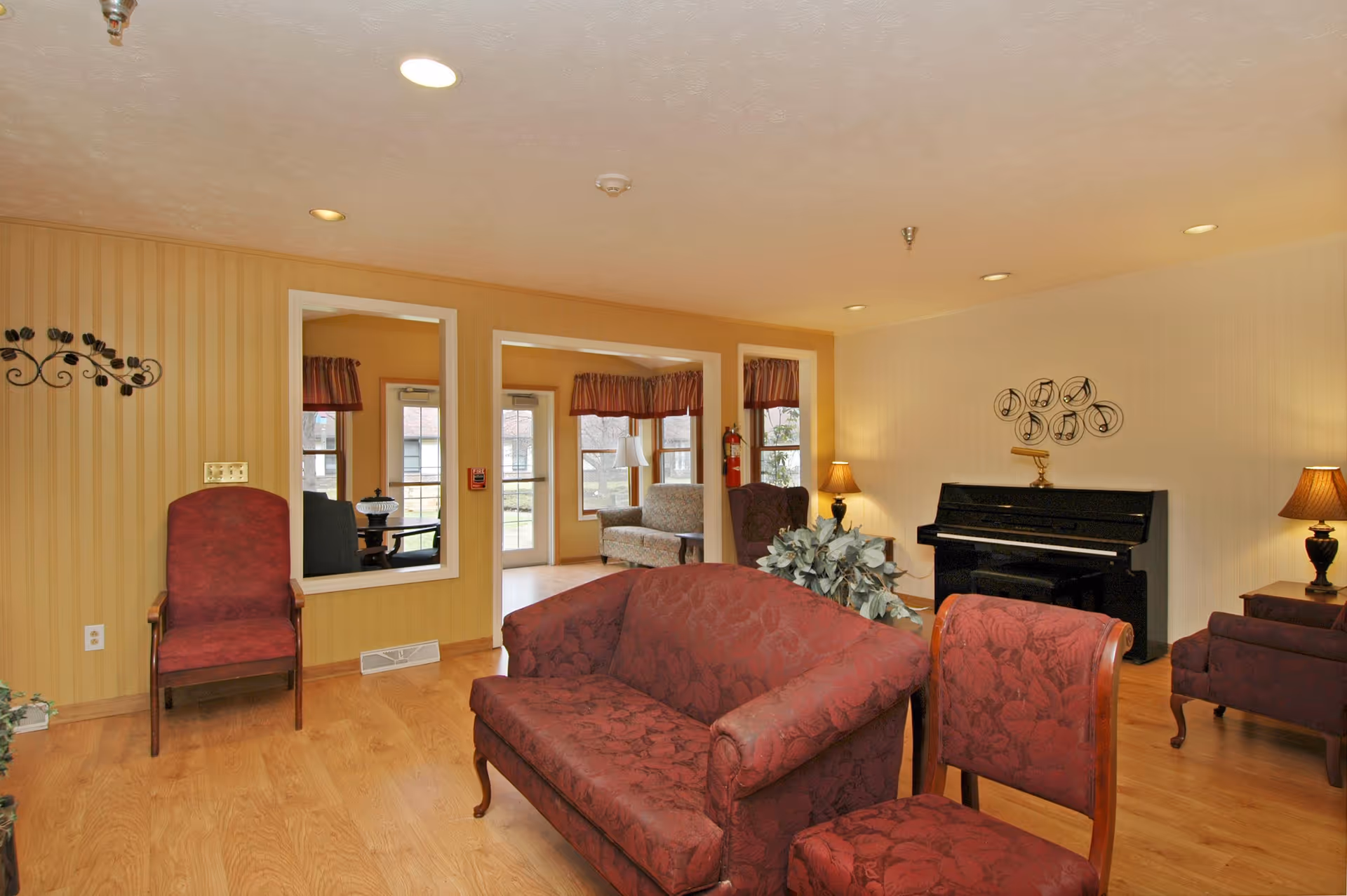 A cozy living room area with wooden flooring and beige striped wallpaper. The room features a red patterned loveseat and matching chair, a black upright piano against the wall with musical note decorations above it, two table lamps with warm lighting, and a large mirror reflecting another seating area with windows and a door leading outside.