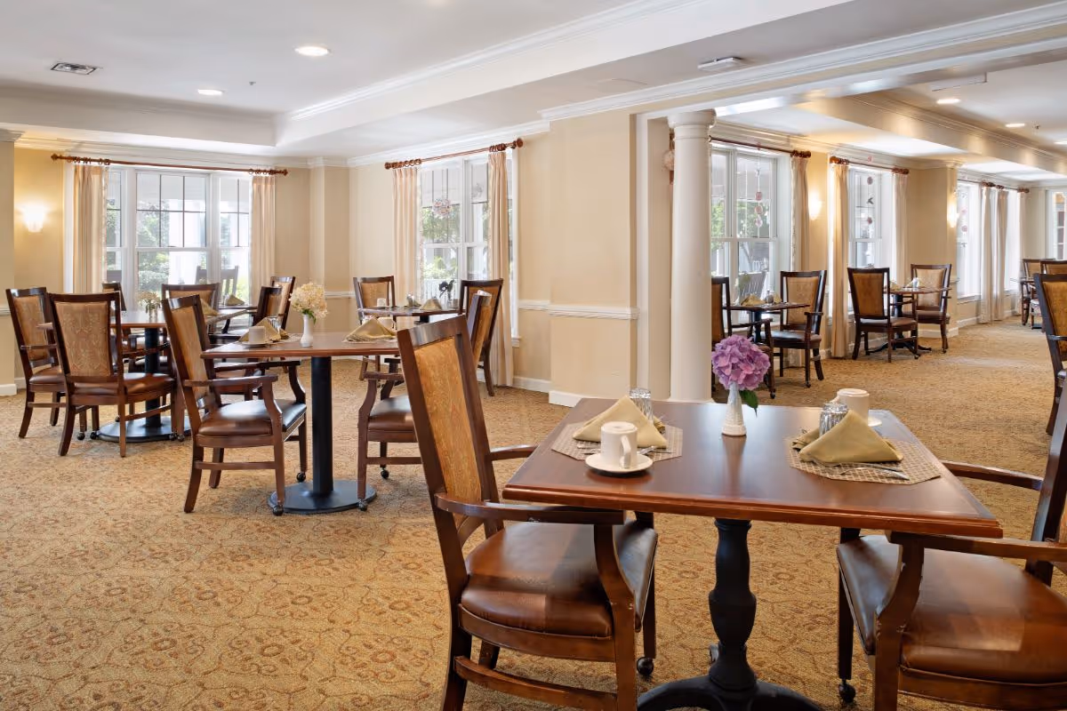A bright and spacious dining room with multiple wooden tables and chairs arranged neatly. Each table is set with beige napkins, cups, and small flower vases. Large windows with light curtains allow natural light to fill the room, and the carpet has a patterned design. The room features cream-colored walls and decorative columns.