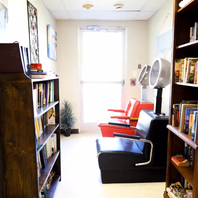 Interior common room with bookshelves and salon-style chairs with hooded hair dryers facing a bright exit door.