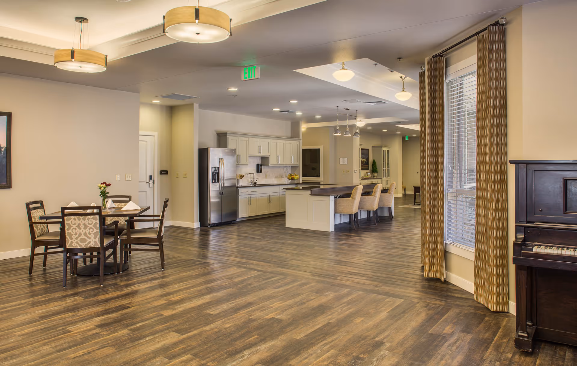 Spacious communal dining and kitchen area with hardwood floors, a dining table, island seating, and a piano by the window.