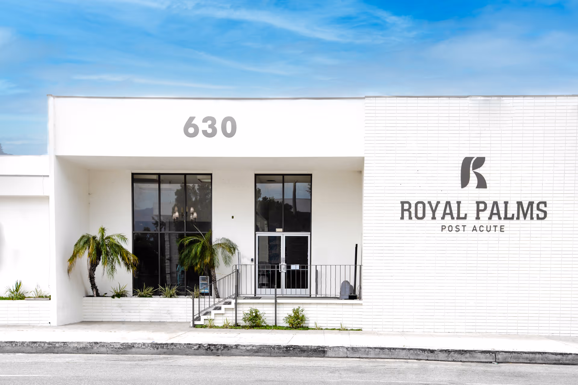 Front exterior view of the Royal Palms Post Acute facility building with large windows, a glass door entrance, and the address number 630 displayed above. There are small palm plants in front of the building and a clear blue sky above.