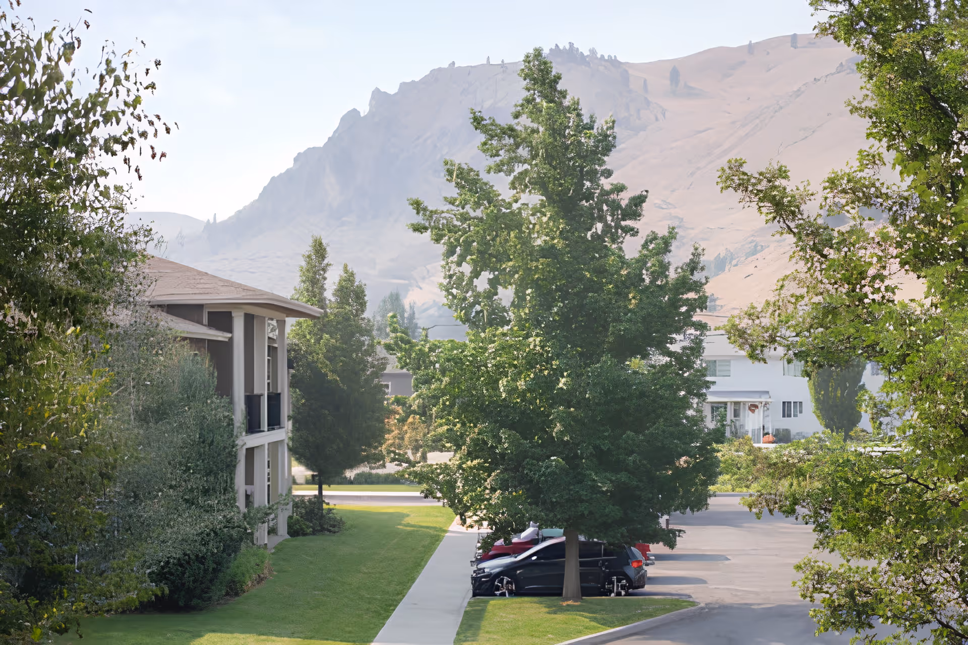 Outdoor view of Wenatchee Senior Living by Cogir showing a parking area with several cars partially shaded by large green trees, a sidewalk, and a building on the left side. In the background, there are dry, rocky hills under a clear sky.