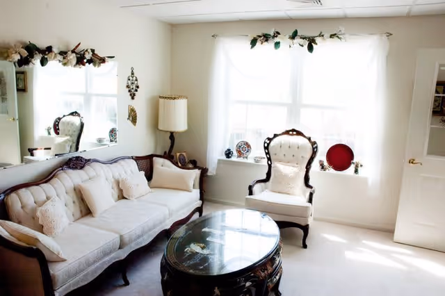 A bright living room with a vintage style featuring a cream-colored tufted sofa with multiple pillows, a matching armchair, a round glass-top wooden coffee table, a floor lamp, and a large window with sheer white curtains decorated with a floral garland.