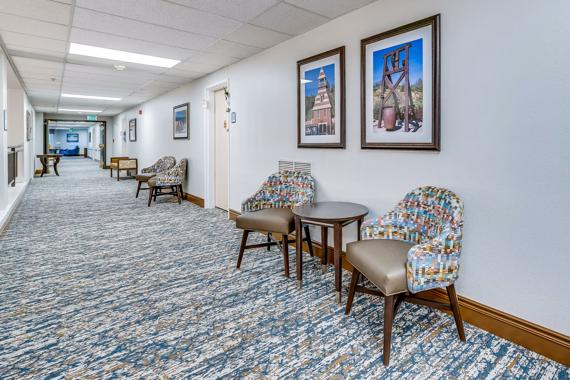 A long, well-lit hallway in a senior living facility with patterned carpet and white walls. Along the right wall, there are colorful upholstered chairs with a small round table between them, and framed pictures hanging above. Additional seating and tables are visible further down the hallway.