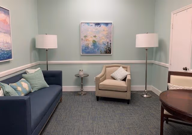 A cozy living room with a blue sofa adorned with multiple cushions, a beige armchair with a patterned pillow, two tall floor lamps with white shades on either side, a small round side table between the sofa and armchair, and a round wooden table with chairs partially visible. The walls are painted light green with white trim, and two framed paintings hang on the walls.