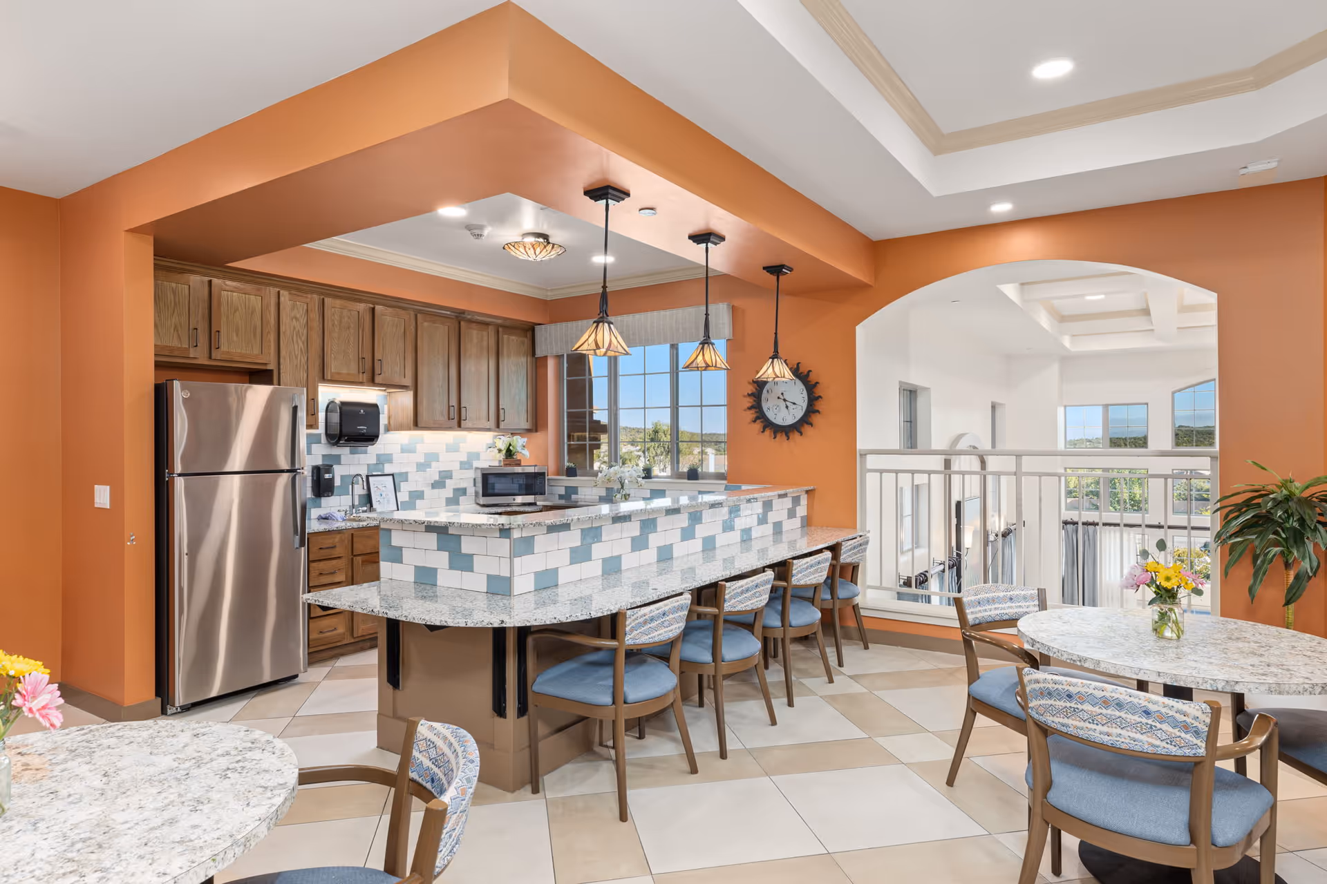 A bright and spacious kitchen and dining area with orange walls and a tiled floor. The kitchen features wooden cabinets, a stainless steel refrigerator, a microwave, and a tiled backsplash in white and blue. There is a granite countertop with bar seating and pendant lights hanging above. The dining area has round tables with floral centerpieces and cushioned chairs. Large windows provide a view of greenery outside.