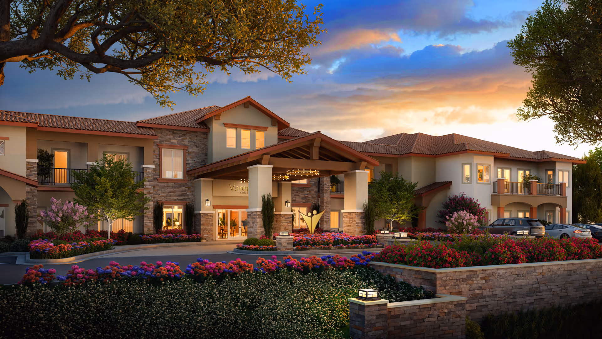 Front exterior of a two-story senior living building with a covered entrance, landscaped flowerbeds and a sunset sky.
