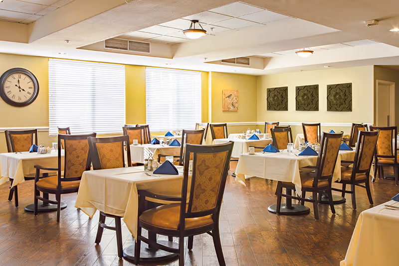 Well-lit dining room with multiple tables covered in white tablecloths, blue folded napkins, and wooden chairs.