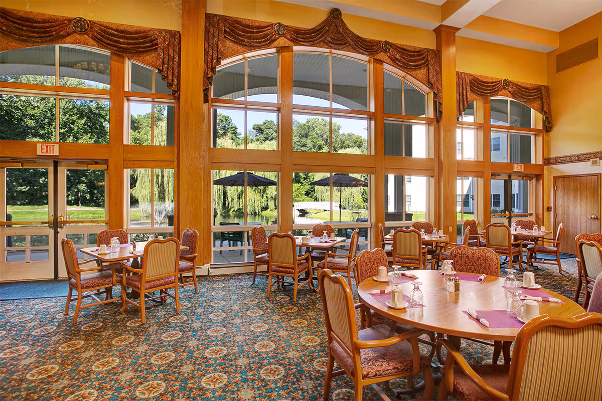 Sunlit dining room with round wooden tables and upholstered chairs set near large floor-to-ceiling windows overlooking a landscaped lawn and pond.