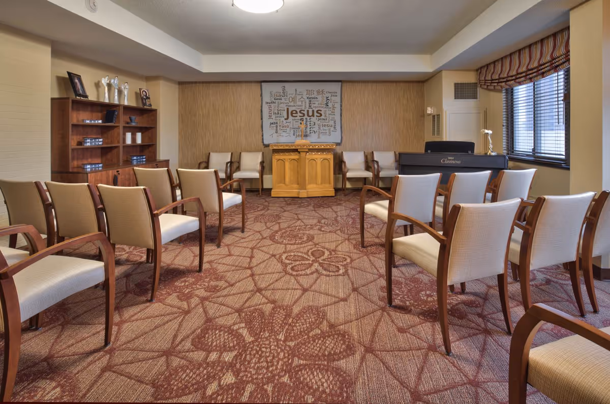A small chapel or prayer room with rows of beige cushioned chairs with wooden arms arranged facing a wooden podium with a cross. Behind the podium is a wall hanging with the word 'Jesus' and other religious words in various languages. To the right is a black electronic piano near a window with striped curtains. On the left side, there is a wooden bookshelf with decorative items and framed photos. The carpet has a floral pattern in shades of red and beige.
