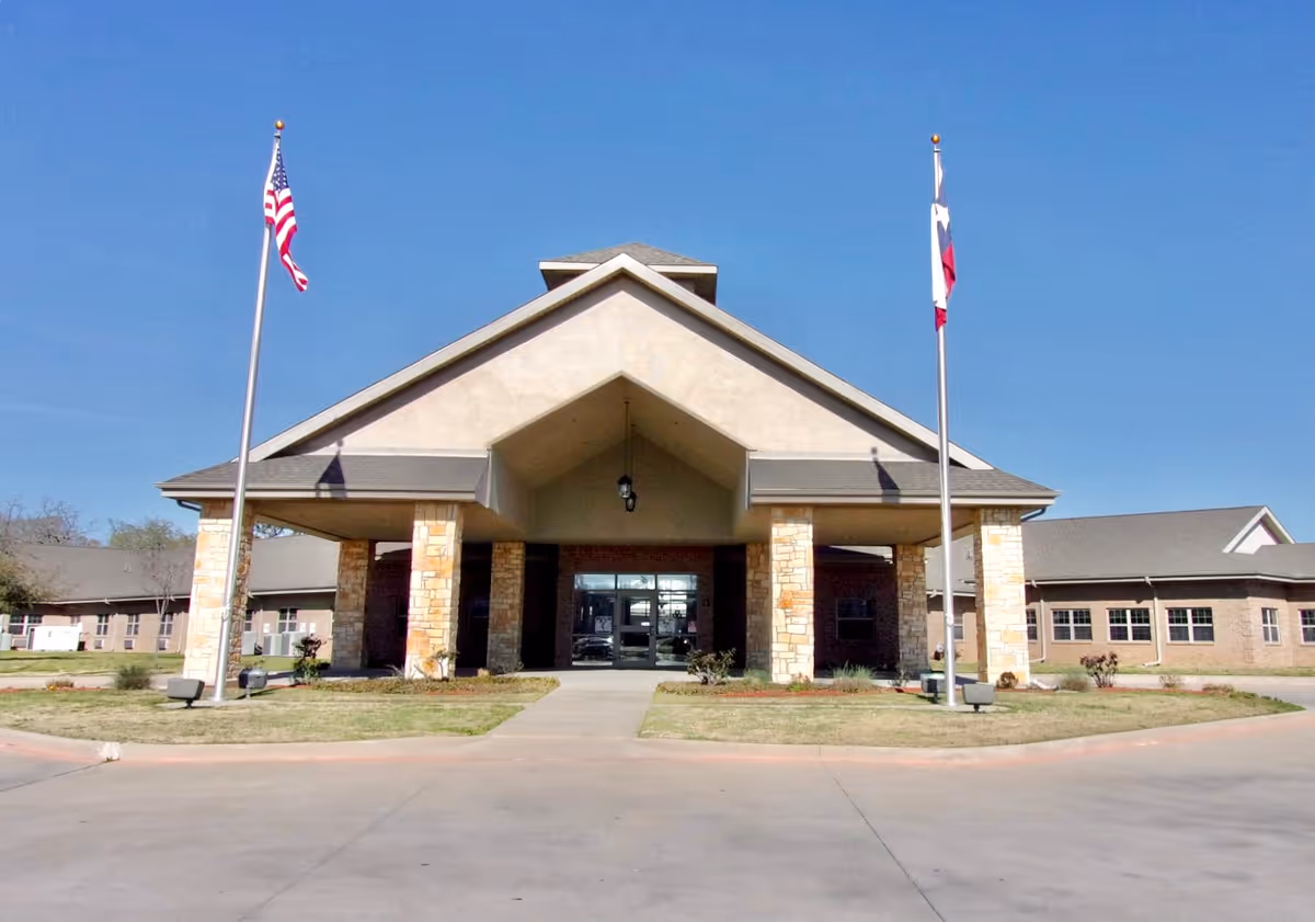 Front exterior view of The Atrium of Bellmead building with a covered entrance supported by stone pillars, two flagpoles with the American and Texas flags, and a clear blue sky.