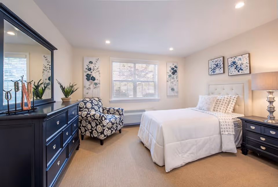 A bright and cozy bedroom with a white bed, patterned armchair, dark blue dresser with a large mirror, and matching nightstand with a lamp. The walls are decorated with floral artwork, and a window with blinds lets in natural light.