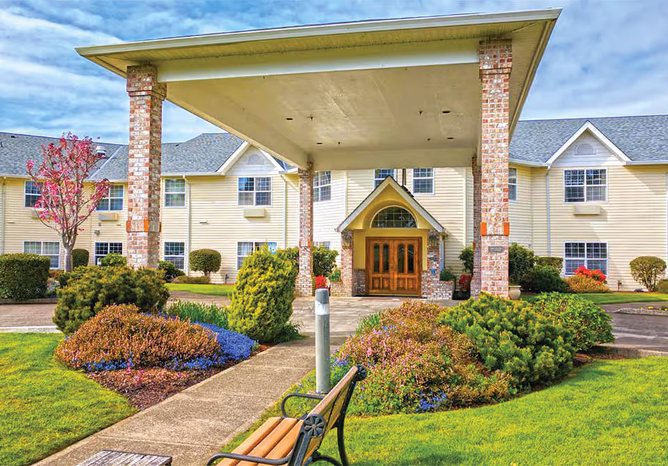 Front exterior view of Lakeview Senior Living building with a covered entrance supported by brick pillars, surrounded by well-maintained landscaping including bushes, flowers, and a tree. A bench is visible near the walkway leading to the entrance.