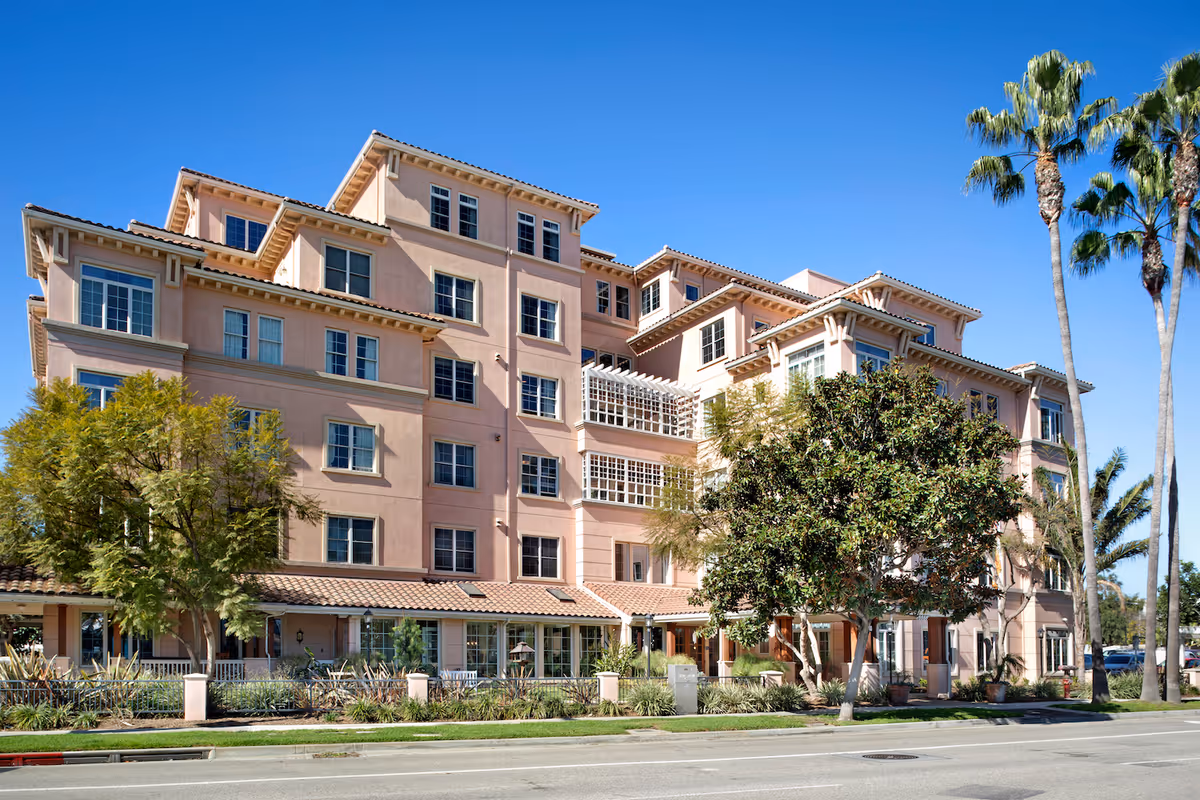 Exterior view of a multi-story senior living facility building with beige stucco walls, multiple windows, and a tiled roof. The building is surrounded by trees and landscaping, with a clear blue sky overhead.