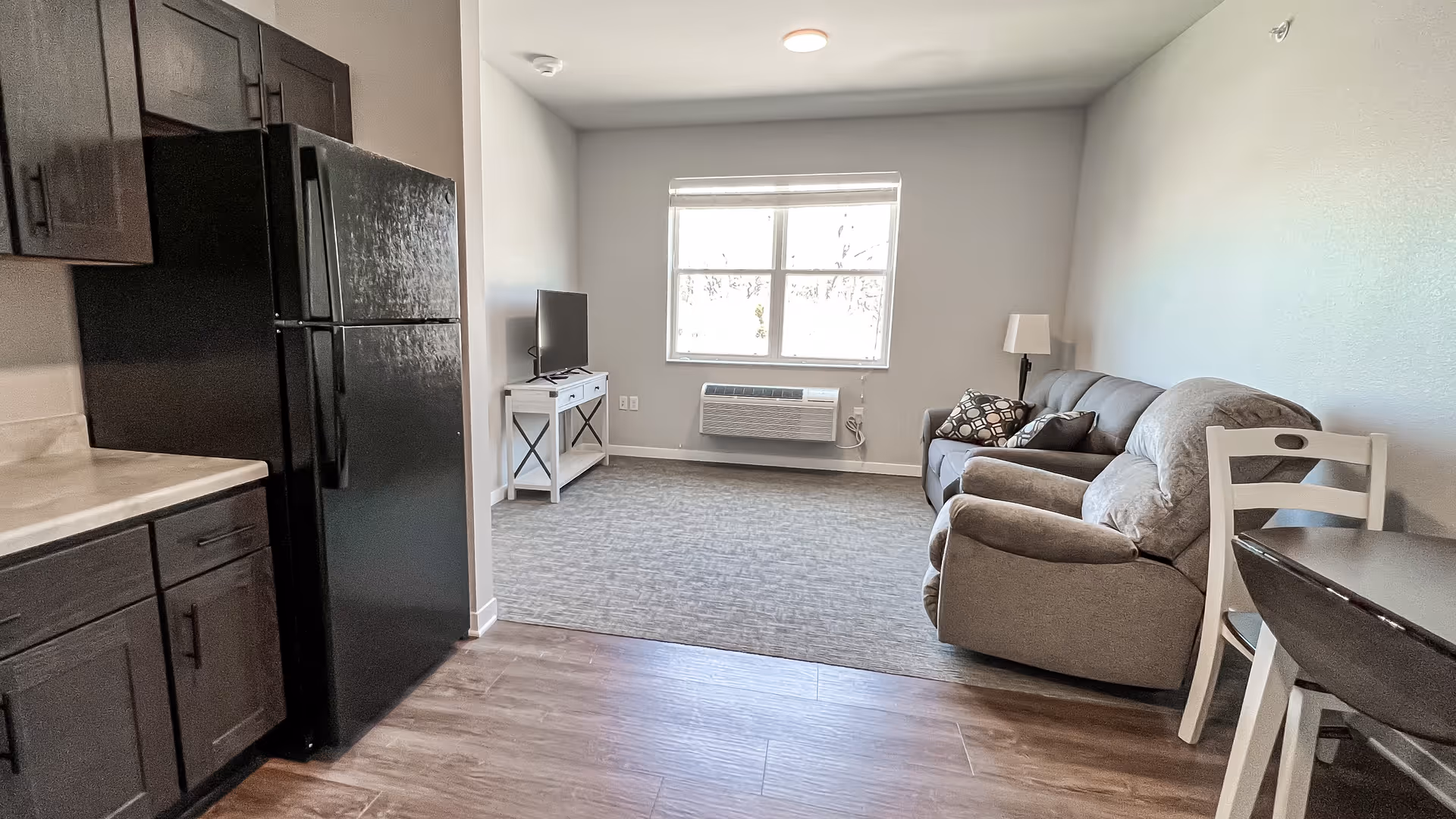 Open-plan apartment living area with a kitchen and black refrigerator on the left and a couch, TV stand and window on the right.