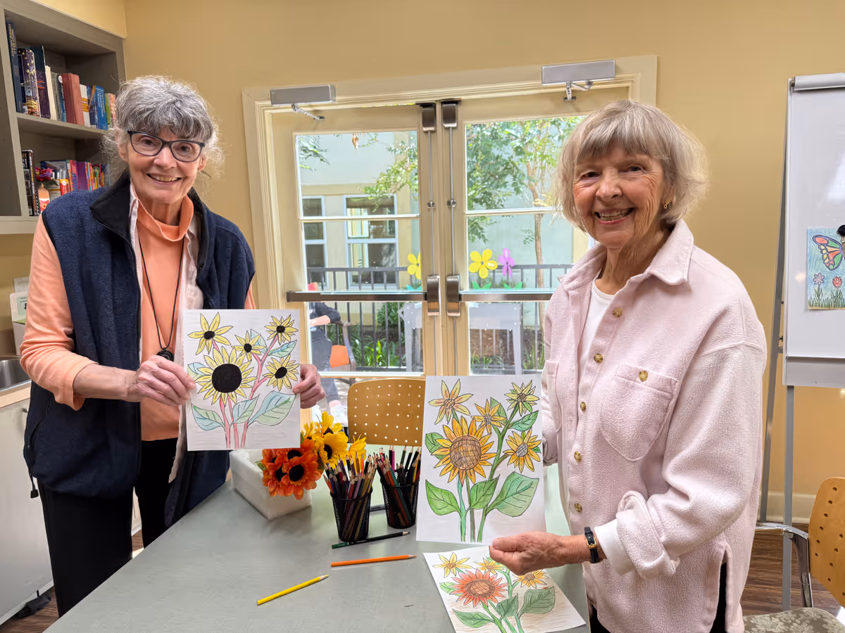 Two elderly women standing indoors at a table, smiling and holding up their colorful drawings of sunflowers. The table has colored pencils and a vase with artificial sunflowers. Behind them is a glass door with flower decorations and a whiteboard with a butterfly drawing.