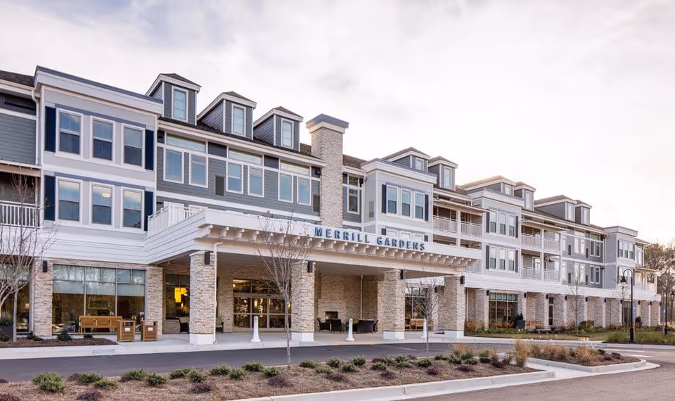 Exterior view of a multi-story senior living facility named Merrill Gardens at Woodstock, featuring a covered entrance with stone pillars, multiple windows, and balconies. The building is surrounded by landscaped grounds and a driveway.