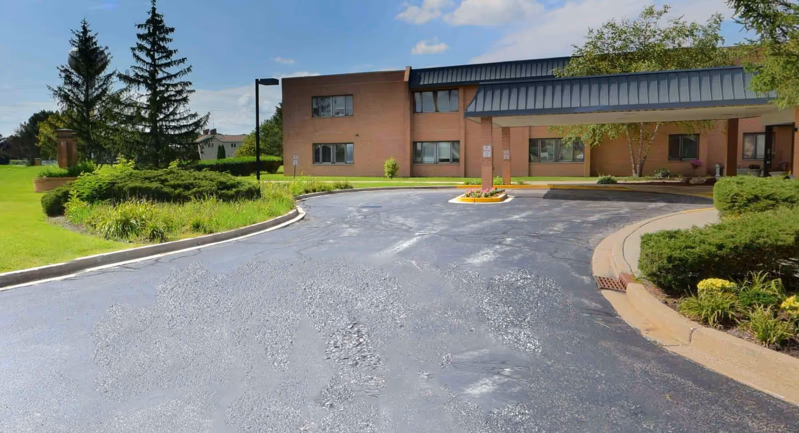 Exterior view of St James Manor & Villas showing a brick building with multiple windows, a covered entrance with a dark metal roof, a curved driveway, and landscaped greenery including bushes, trees, and grass under a partly cloudy sky.