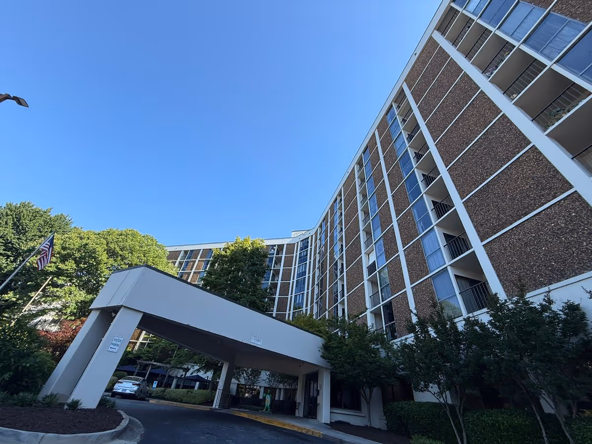 Low-angle view of the Campbell-Stone Sandy Springs multi-story building with a covered drop-off entrance, trees, and a clear blue sky.