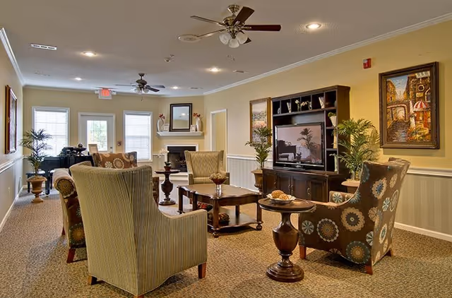 A cozy living room area in Montgomery Gardens featuring several upholstered armchairs arranged around wooden coffee tables. The room has a carpeted floor, ceiling fans, a TV on a wooden entertainment center, framed artwork on the walls, and potted plants adding greenery. Natural light enters through windows and a glass door at the far end.