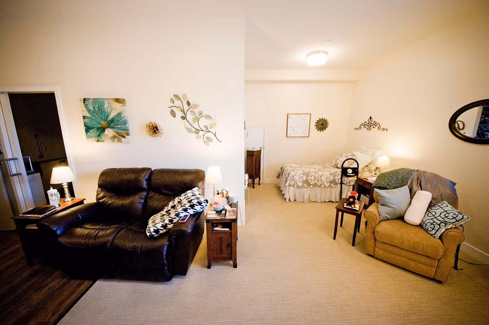 A cozy assisted living room featuring a dark brown leather loveseat with a patterned pillow, a wooden side table with a lamp and tissues, and a beige armchair with multiple pillows and a blanket. In the background, there is a bed with a floral bedspread, a small wooden nightstand with a lamp, and decorative wall art including a large flower painting and metal leaf designs. The room has beige carpet and soft lighting.