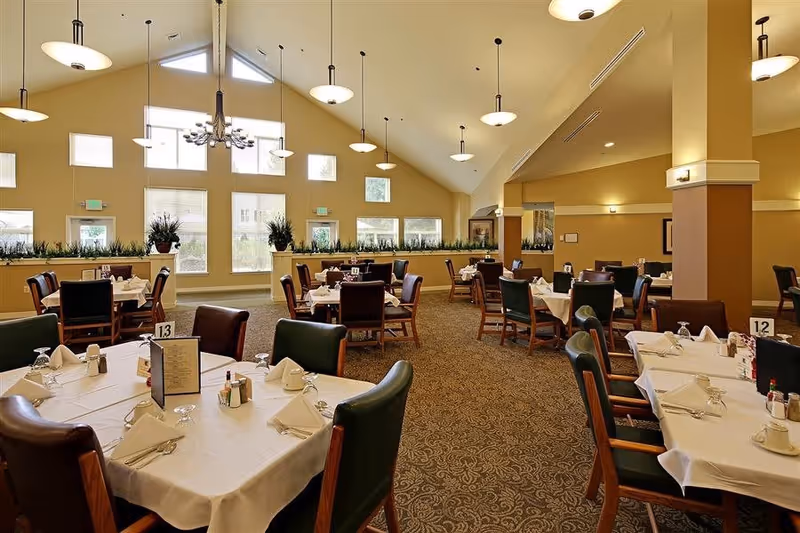 A spacious dining room with multiple tables covered in white tablecloths, each set with napkins, glasses, and menus. The room features high vaulted ceilings with hanging light fixtures, large windows letting in natural light, and potted plants along the window ledge.