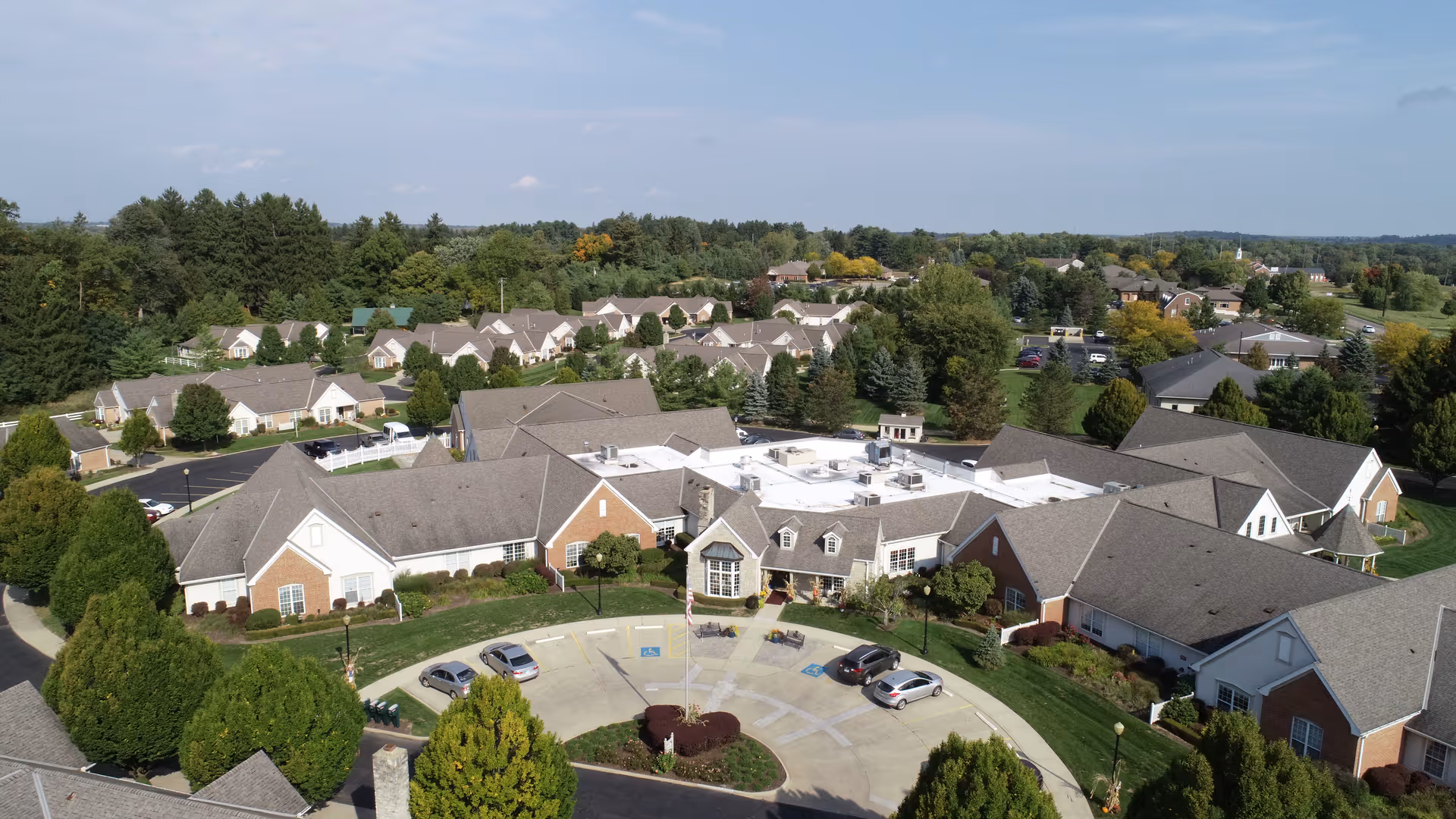 Aerial view of Wedgewood Estates senior living facility showing multiple connected buildings with gray roofs and brick accents, surrounded by trees and greenery. There is a circular driveway with parked cars and a flagpole in the center. Residential houses and more greenery are visible in the background under a partly cloudy sky.